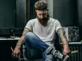Mężczyzna z brodą i tatuażem na biceps siedzi w fotelu, trzymając nożyczki.