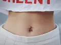 Brzuszek z kolczykiem w pępku. Czy dziurka w pępku zarasta? Delikatna skóra, białe spodenki i crop top z czerwonym napisem.