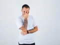 Mężczyzna w białej koszulce zakrywa twarz dłonią, odsłaniając rękę z wypukłym tatuażem.