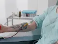 Osoba z tatuażem na ramieniu oddaje krew, ściskając czerwoną piłeczkę. W tle stoją probówki.