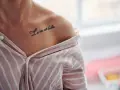 Delikatny tatuaż napis na obojczyku: "La vie est belle". Kobieta w pasiastym, rozpiętym dekoltem ubraniu.