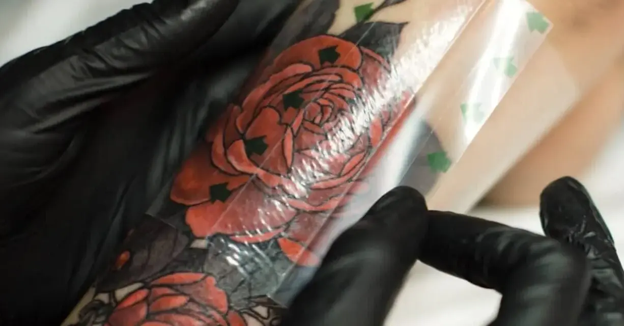 Ręce w czarnych rękawiczkach zdejmują folię ochronną z tatuażu z czerwonymi różami. To kluczowy etap, jak zdjąć second skina, by tatuaż prawidłowo się goił.