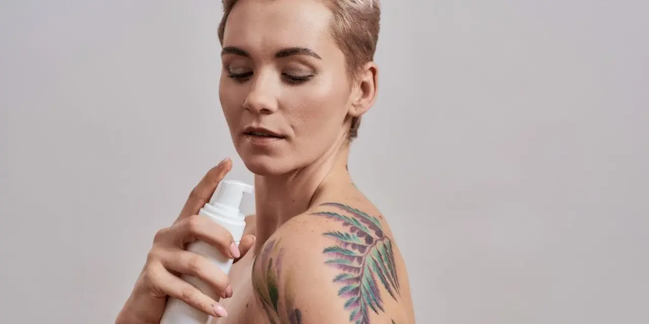 Kobieta z kolorowym tatuażem na ramieniu aplikuje mgiełki do pielęgnacji tatuażu.