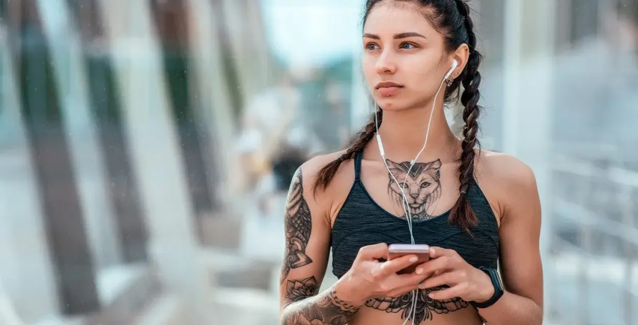 Kobieta z warkoczami i tatuażem na klacie, słuchająca muzyki na telefonie.