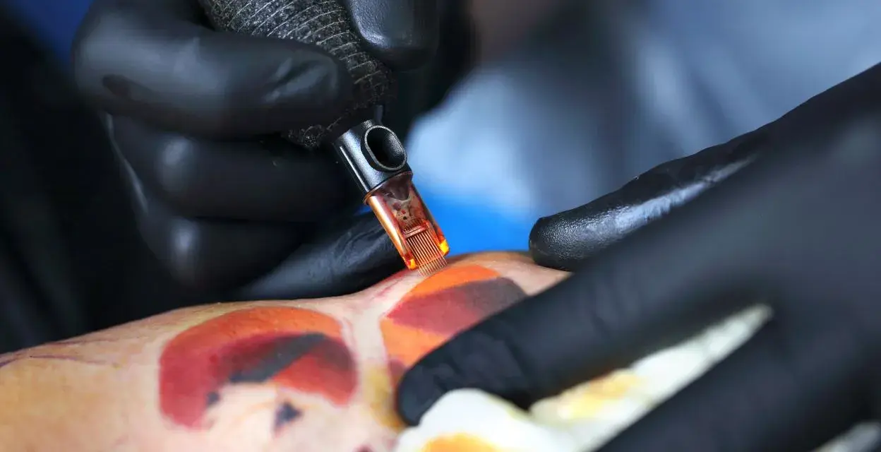 Artysta w czarnych rękawiczkach tworzy kolorowy tatuaż. Pielęgnacja świeżego tatuażu jest kluczowa, a pytanie "ile razy dziennie myc tatuaz" jest ważne dla jego prawidłowego gojenia.