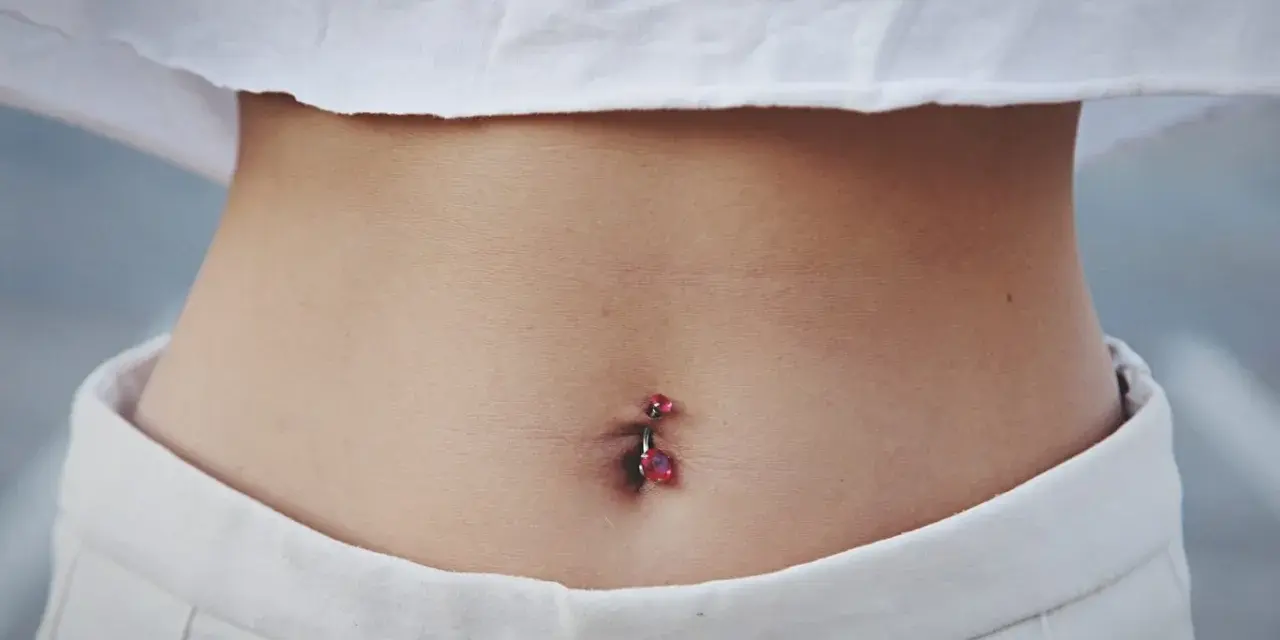 Brzuszek z kolczykiem w pępku. Czy dziurka w pępku zarasta? Delikatna skóra, białe spodenki i crop top z czerwonym napisem.