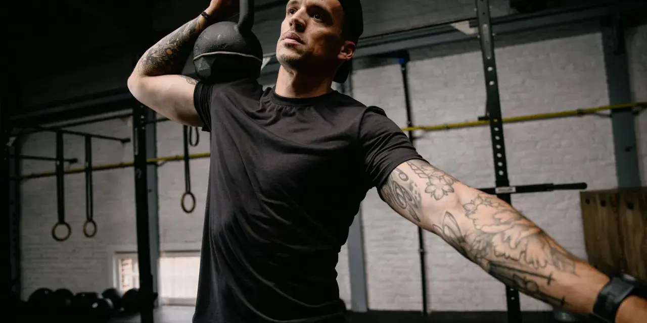 Mężczyzna z małym tatuażem na ramieniu wykonuje ćwiczenie z kettlebell. Intensywny trening w siłowni.