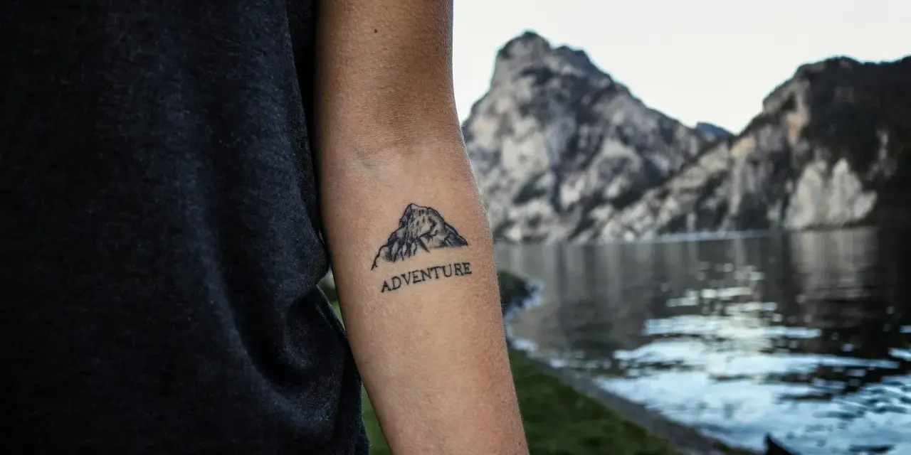 Tatuaż góry na przedramieniu z napisem "ADVENTURE". Symbolizuje miłość do podróży i odkrywania nowych miejsc.