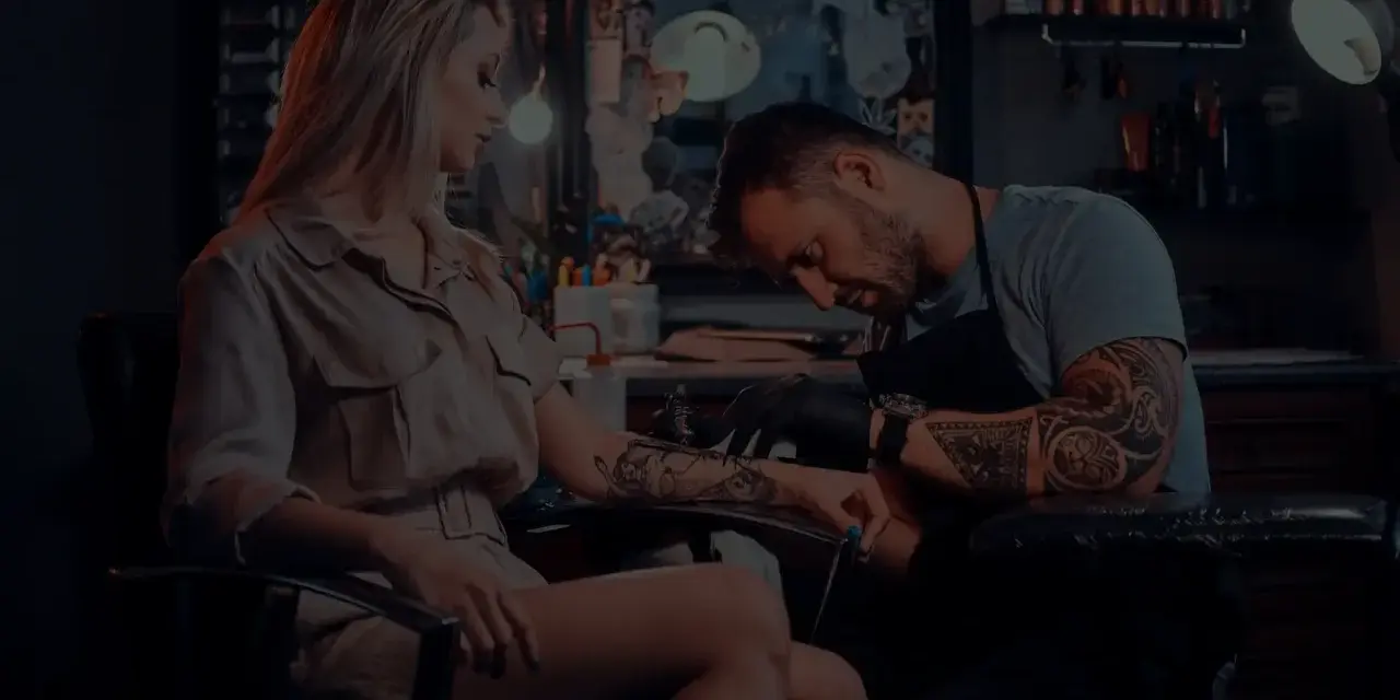 Artysta tworzy tatuaż opaska na nodze kobiety. W tle widać studio tatuażu.