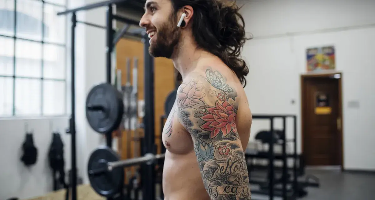 Uśmiechnięty mężczyzna z tatuażem na ramieniu w siłowni. Czy po tatuazu mozna cwiczyc? Tak, jeśli jest zagojony!