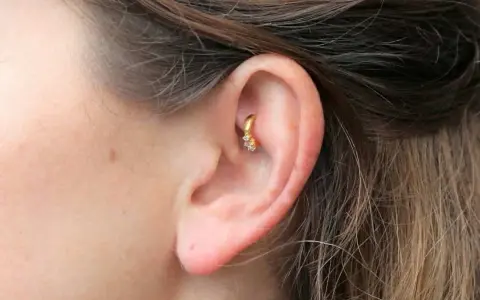 Złoty kolczyk w uchu, ozdobiony cyrkoniami, w trakcie gojenia. Rook piercing wygląda pięknie.