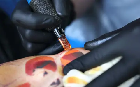 Artysta w czarnych rękawiczkach tworzy kolorowy tatuaż. Pielęgnacja świeżego tatuażu jest kluczowa, a pytanie "ile razy dziennie myc tatuaz" jest ważne dla jego prawidłowego gojenia.