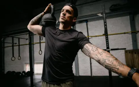 Mężczyzna z małym tatuażem na ramieniu wykonuje ćwiczenie z kettlebell. Intensywny trening w siłowni.