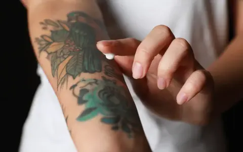 Dłoń nakłada krem na rękę z kolorowym tatuażem, który zaczyna się łuszczyć.