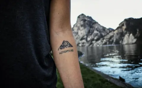 Tatuaż góry na przedramieniu z napisem "ADVENTURE". Symbolizuje miłość do podróży i odkrywania nowych miejsc.