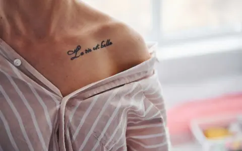 Delikatny tatuaż napis na obojczyku: "La vie est belle". Kobieta w pasiastym, rozpiętym dekoltem ubraniu.