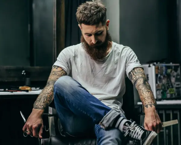 Mężczyzna z brodą i tatuażem na biceps siedzi w fotelu, trzymając nożyczki.