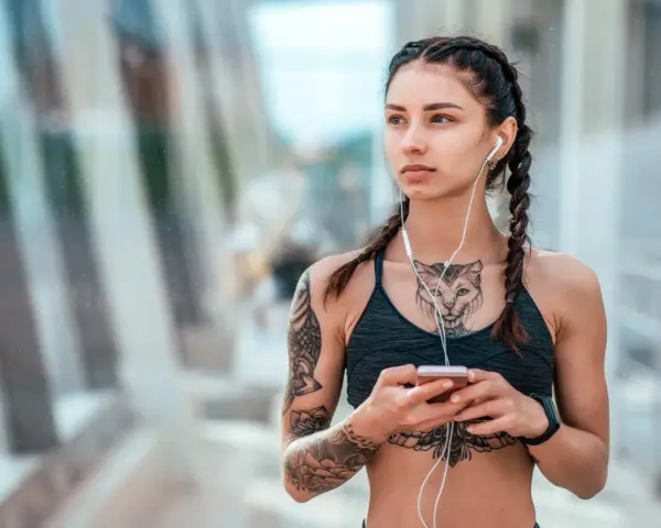 Kobieta z warkoczami i tatuażem na klacie, słuchająca muzyki na telefonie.