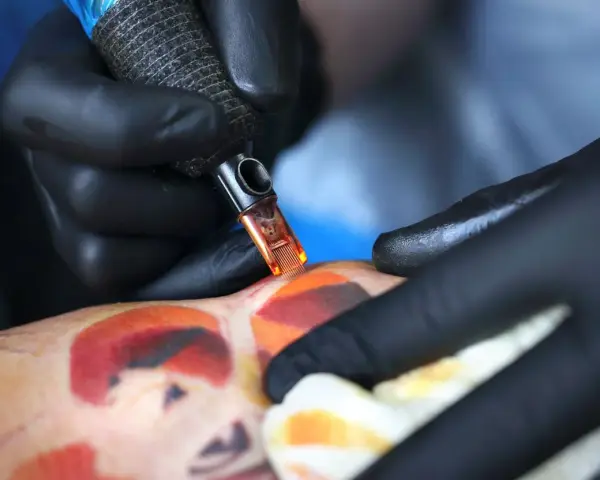 Artysta w czarnych rękawiczkach tworzy kolorowy tatuaż. Pielęgnacja świeżego tatuażu jest kluczowa, a pytanie "ile razy dziennie myc tatuaz" jest ważne dla jego prawidłowego gojenia.