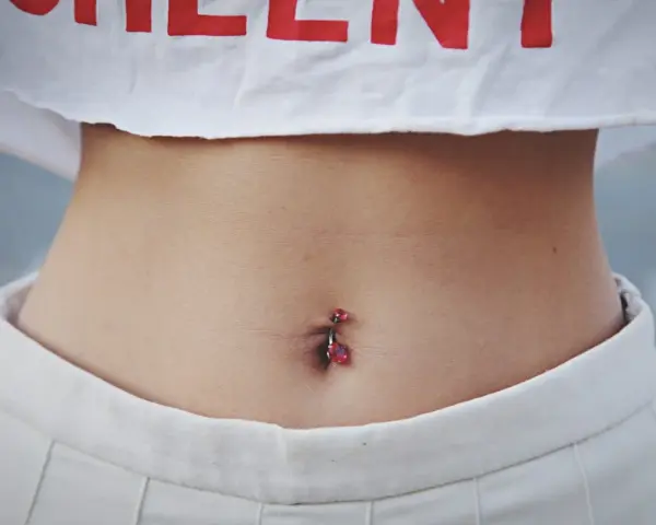 Brzuszek z kolczykiem w pępku. Czy dziurka w pępku zarasta? Delikatna skóra, białe spodenki i crop top z czerwonym napisem.
