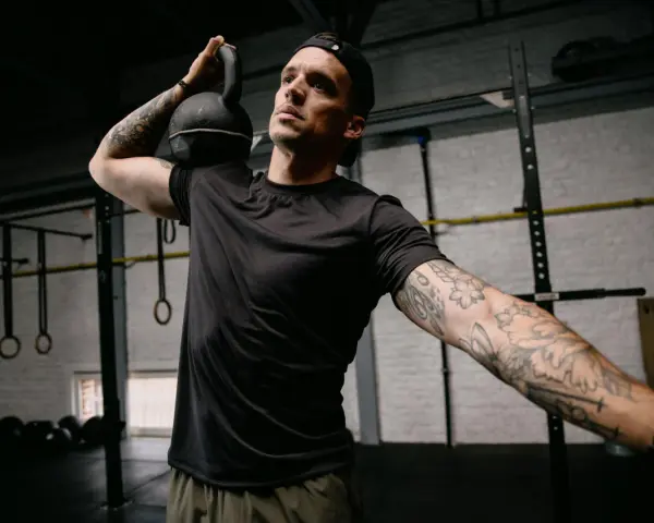 Mężczyzna z małym tatuażem na ramieniu wykonuje ćwiczenie z kettlebell. Intensywny trening w siłowni.