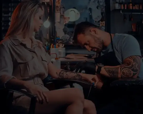 Artysta tworzy tatuaż opaska na nodze kobiety. W tle widać studio tatuażu.