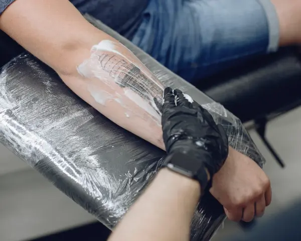 Nowy tatuaż na przedramieniu, artysta w rękawiczce nakłada piankę. Pytanie: nowy tatuaż jak długo nosić folię?