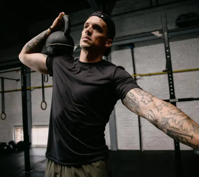 Mężczyzna z małym tatuażem na ramieniu wykonuje ćwiczenie z kettlebell. Intensywny trening w siłowni.