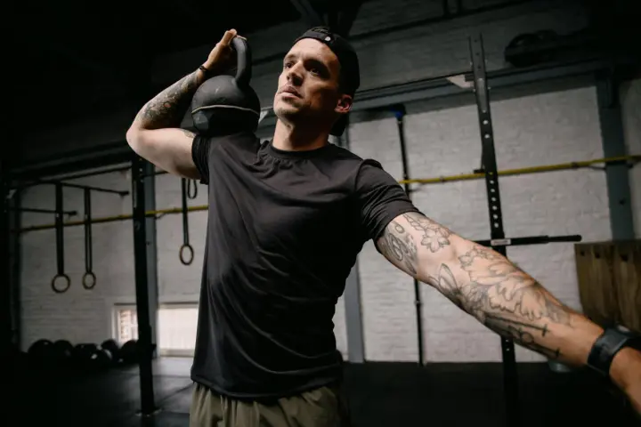 Mężczyzna z małym tatuażem na ramieniu wykonuje ćwiczenie z kettlebell. Intensywny trening w siłowni.