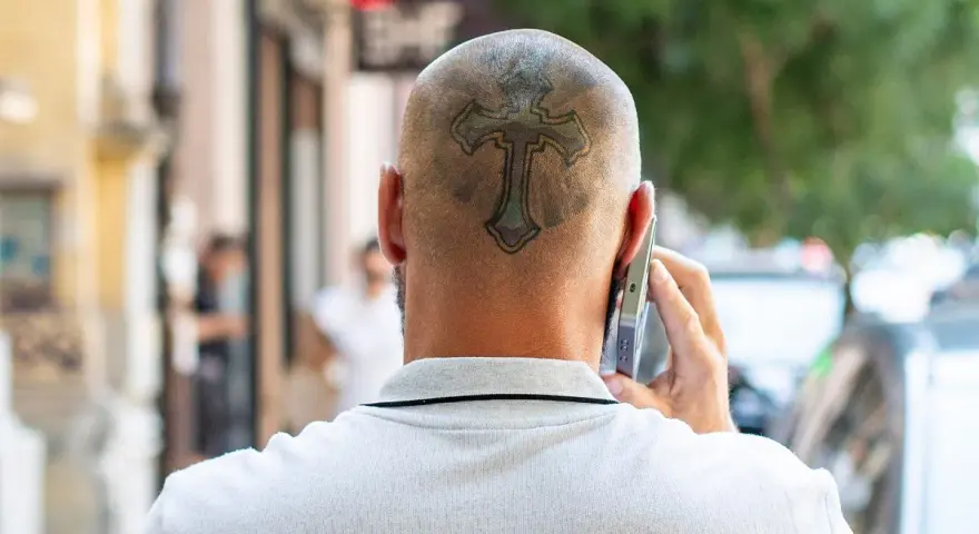 Mężczyzna rozmawia przez telefon, a na jego ogolonej głowie widoczny jest tatuaż krzyża.