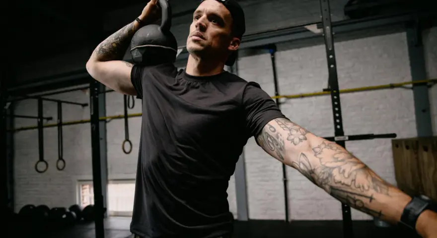 Mężczyzna z małym tatuażem na ramieniu wykonuje ćwiczenie z kettlebell. Intensywny trening w siłowni.