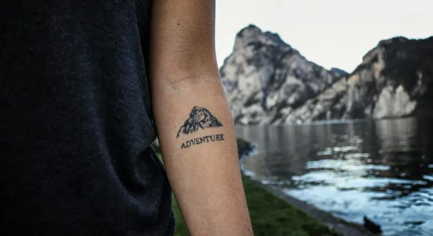 Tatuaż góry na przedramieniu z napisem "ADVENTURE". Symbolizuje miłość do podróży i odkrywania nowych miejsc.