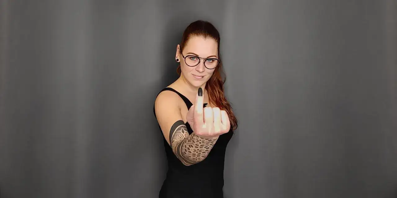 Kobieta z tatuażem na ręce pokazuje środkowy palec, z wyrazem twarzy sugerującym, że nawet tatuaż podczas okresu nie powstrzyma jej przed wyrażeniem siebie.