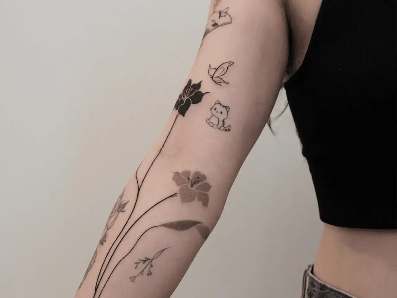 Delikatny tatuaż na boku ręki: kwiaty, motyl i kotek.