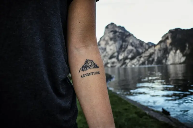 Tatuaż góry na przedramieniu z napisem "ADVENTURE". Symbolizuje miłość do podróży i odkrywania nowych miejsc.